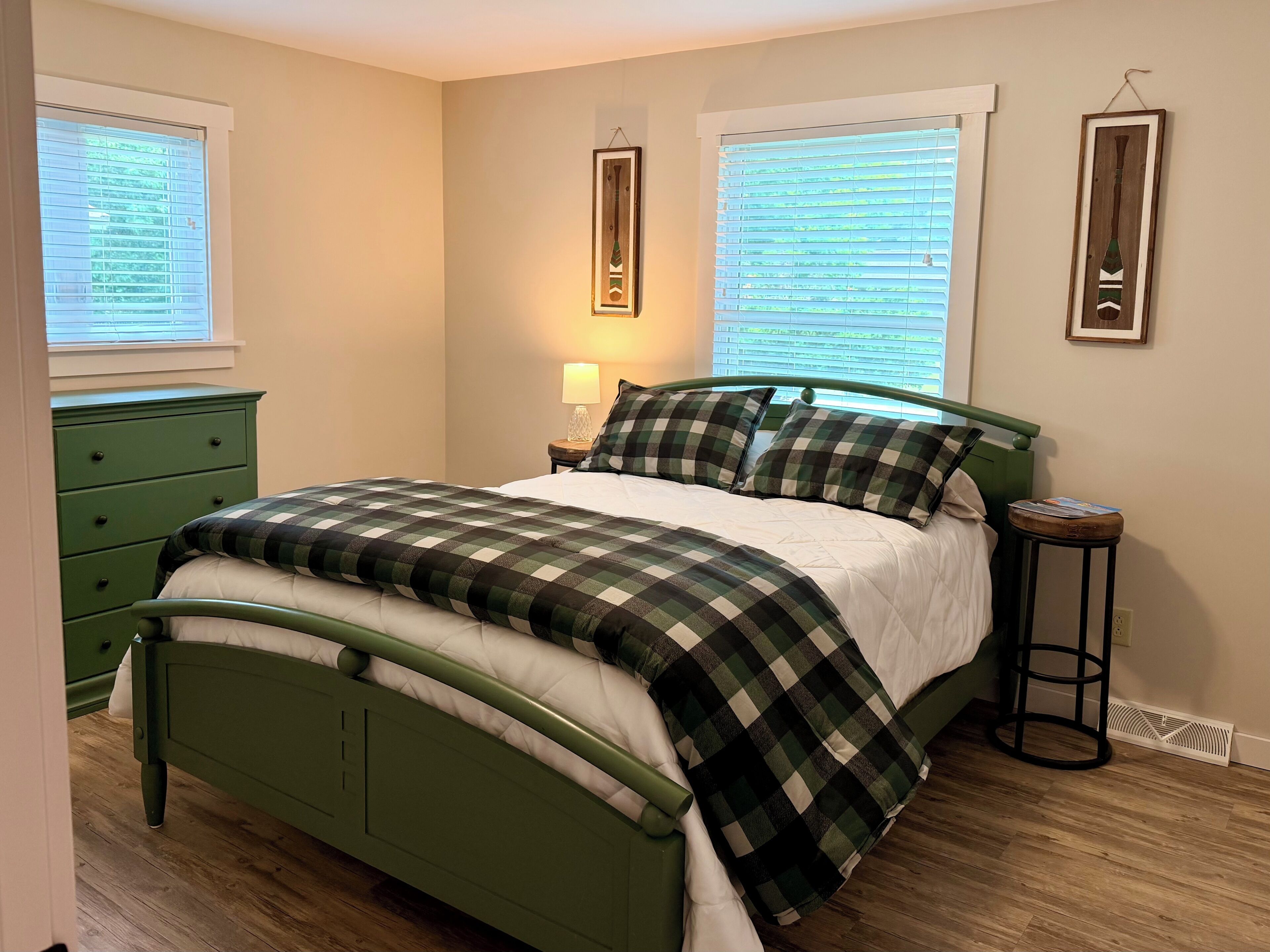 A bedroom with a green metal bed frame, a checkered bedspread, two bedside tables with lamps, and a dresser.