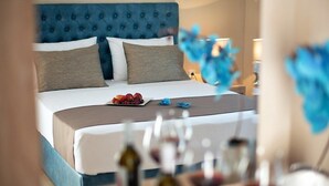 Ein Hotelzimmer mit Bett, Kissen, einem Nachttisch mit Obst und einer Flasche sowie einer Vase mit Blumen.