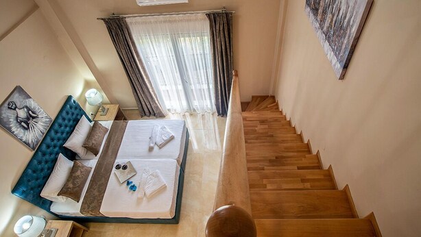 Ein Schlafzimmer mit einem Bett, einem blauen Kopfende, einem Nachttisch mit Lampe, einem Fenster mit Vorhängen und einer Treppe.