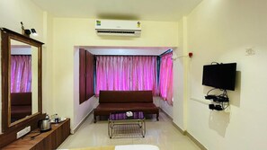 A hotel room with a sofa, a TV, an air conditioner, a mirror, and a window with curtains.