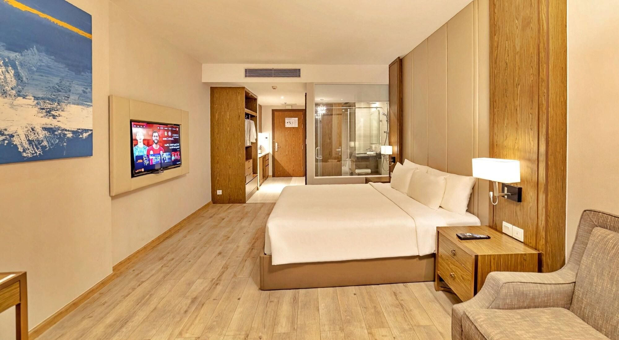 Habitación de hotel con cama, televisor, silla y baño visible a través de una puerta de cristal.