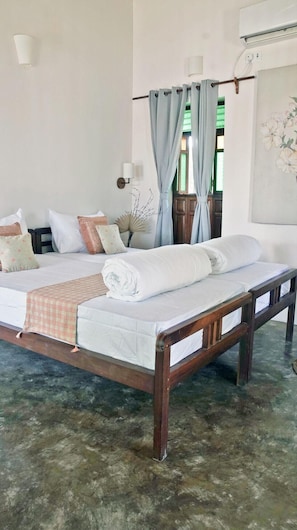 Ein Bett mit Holzrahmen, einer weißen Matratze und einer Decke. Kissen und ein dekorativer Fächer liegen auf dem Bett. Der Raum hat einen Fliesenboden und ein Fenster mit Vorhängen.
