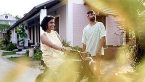 Una persona en bicicleta, otra persona de pie a un lado, y un edificio con porche.