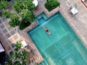 Luftaufnahme eines Schwimmbeckens mit einer Person in einem roten Badeanzug, umgeben von Grünflächen und Steinböden.