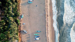 Vista aérea de una playa con varios barcos alineados en la arena, una vista clara de las olas del océano y abundante vegetación a la izquierda.