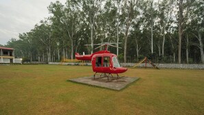 Um helicóptero vermelho sobre um bloco de concreto em um campo gramado, com árvores ao fundo.