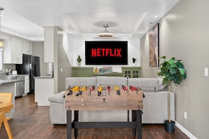 A modern living room with a foosball table, a television displaying 'NETFLIX,' and a kitchen in the background.
