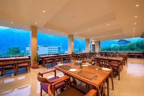 A spacious dining area with multiple tables and chairs, large windows offering a view of the mountains, and a modern ceiling design. - Naturevilas Sarovar Portico (Manali)
