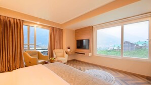 Habitación de hotel con cama, dos sillones, televisor y ventanas amplias con vistas a las montañas.