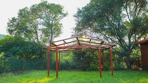 En pergola med gennemsigtigt tag, omgivet af frodig vegetation og træer.