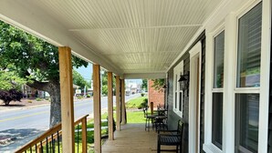 A covered wooden deck with outdoor seating, a railing, and a view of the street and greenery.