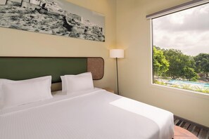 Et hotelværelse med en seng, en lampe og et vindue med udsigt over en pool og grøn bevoksning.