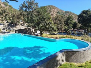A clear blue swimming pool with a curved edge, surrounded by trees and a hilly landscape. - Kumbhalgarh Valley Resort Kumbhalgarh (Kumbhalgarh)