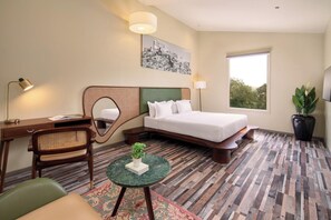 A hotel room with a bed, desk, chair, round table with a plant, a large window, and a painting on the wall. - Kumbhalgarh Valley Resort Kumbhalgarh (Kumbhalgarh)