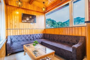 A room with wooden walls, a grey sofa, a glass coffee table, and a TV mounted on the wall. - Hotel Winterfell Mussoorie (Dehradun)