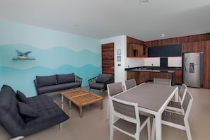 Una cocina moderna con horno y refrigerador integrados, un comedor con mesa de madera y sillas blancas, y una sala de estar con sofá y sillones.