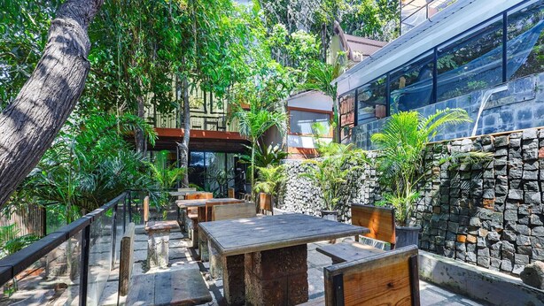 An outdoor seating area with stone tables and wooden chairs, surrounded by lush greenery and a stone wall. - Fabhotel Prime Amazonika Boutique Resort (Arpora)
