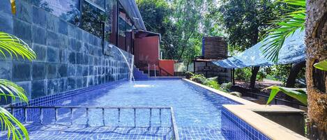 A swimming pool with a tiled edge, surrounded by greenery and a building with large windows.