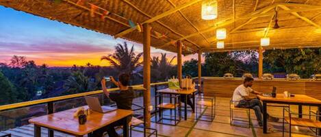 An outdoor dining area with tables and chairs, a thatched roof, and a view of the landscape.