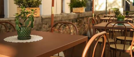 Un espacio al aire libre para comer con mesas y sillas, plantas en macetas y un muro de piedra.