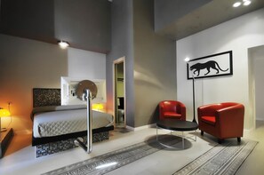Un dormitorio moderno con una cama, dos sillas rojas, una mesita, una lámpara de pie y un cuadro de un gato colgado en la pared.