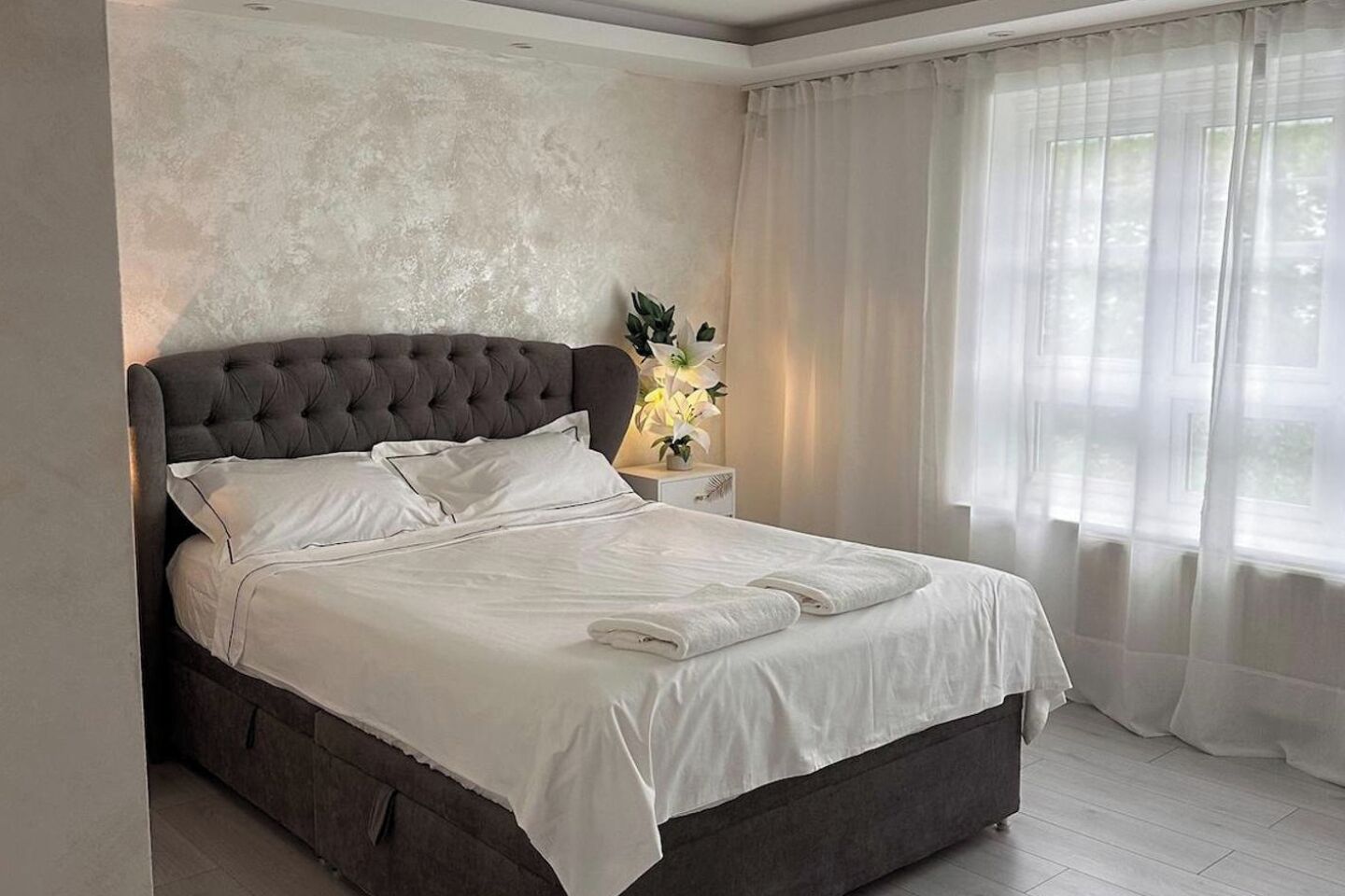 Een slaapkamer met een groot bed, een nachtkastje, een lamp en transparante gordijnen.