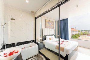 A hotel room with a bathtub, a bed, a large mirror, and a view of buildings. - Elegant Hotel (Xuan Huong - Da Lat)