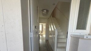 Un pasillo que lleva a una escalera con un candelabro, una puerta con buzón y un ventanal con persianas.