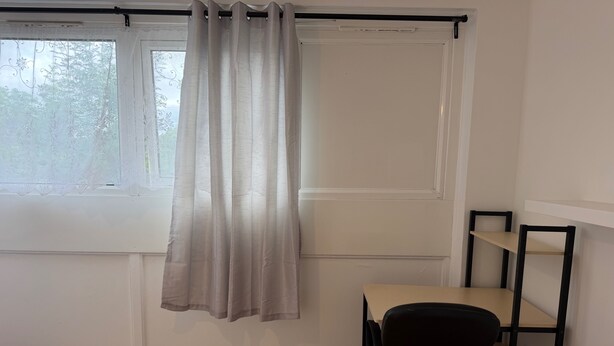 Una habitación con ventana, cortina, escritorio con silla y estantería.