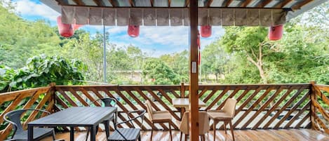 A wooden deck with tables and chairs, a pergola with fabric curtains, and a view of greenery.