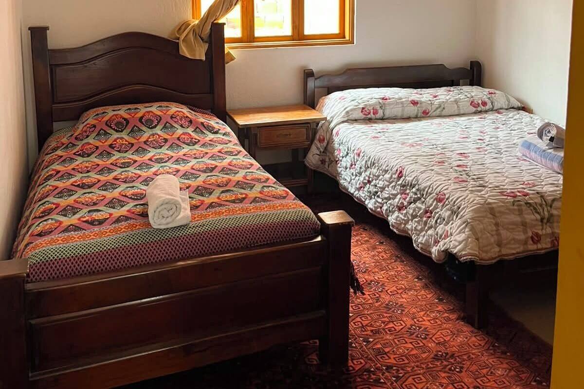 Una habitación con dos camas de madera, una ventana con cortinas, una mesita pequeña y una alfombra estampada.