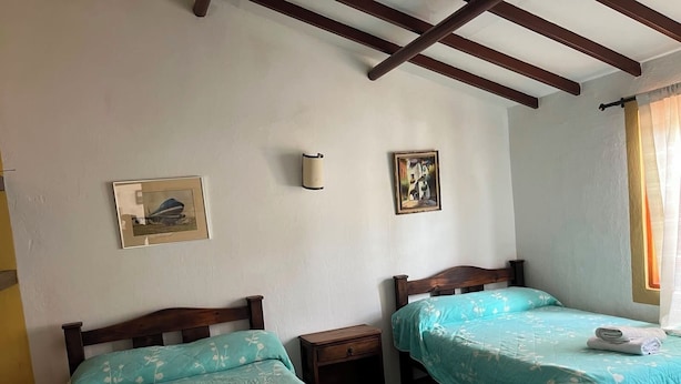 Ein Zimmer mit zwei Betten, beide mit blumigem, blauem Überwurf. Auf einem Bett liegt ein zusammengerolltes Handtuch. Es gibt zwei Nachttische, eine Wandlampe und zwei gerahmte Bilder.