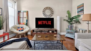 Un salon avec un téléviseur affichant le logo « NETFLIX », un miroir rond, une plante en pot et un fauteuil confortable.