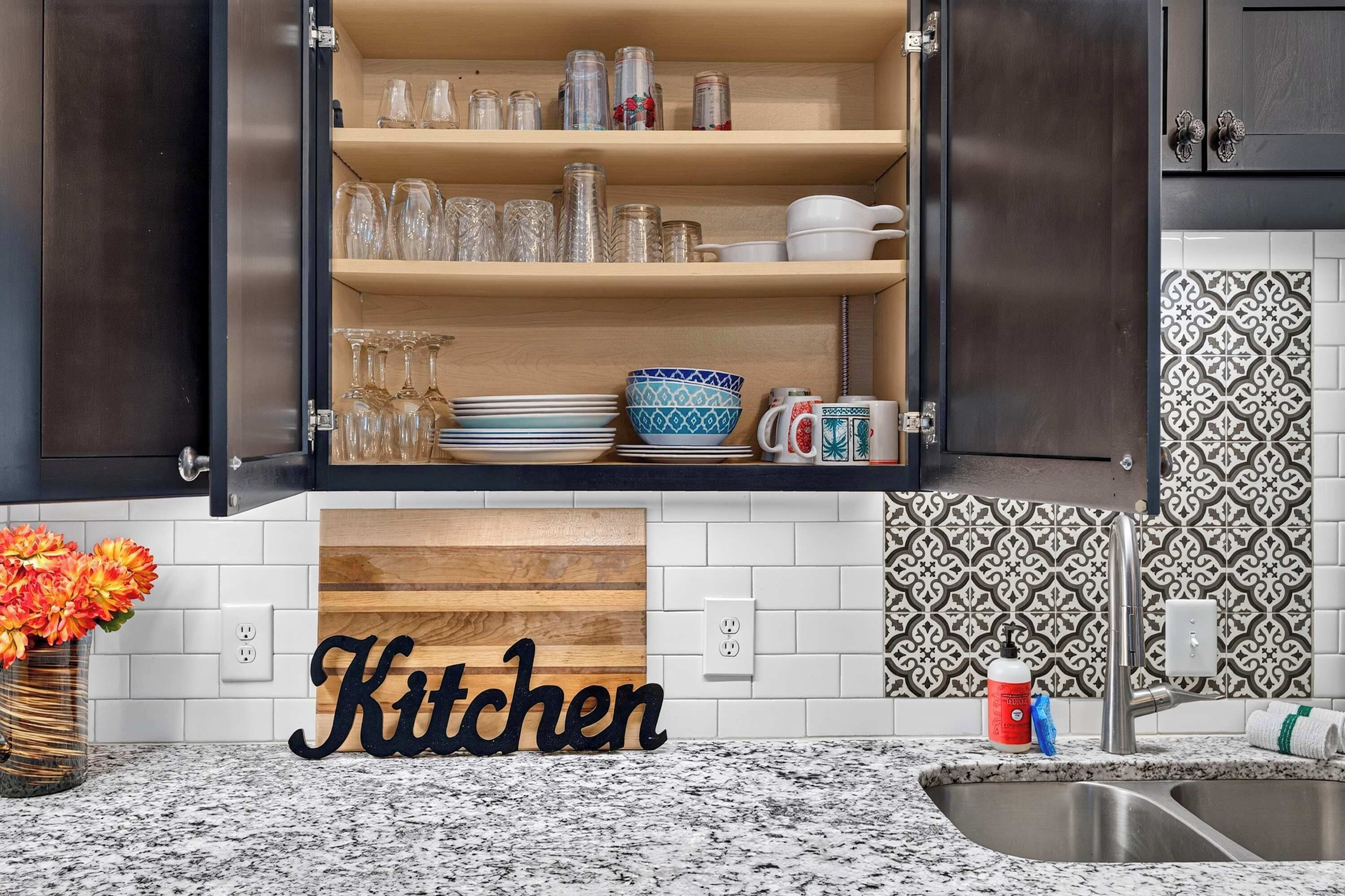 A kitchen with open cabinets displaying glassware, bowls, and plates. A wooden sign with the word 'Kitchen' is on the countertop.