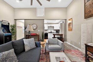 A living room with a couch, armchair, coffee table, and a small table with a lamp. There is a kitchen in the background with cabinets and a clock on the wall. - The Whiskey Stop! A Spacious Highlands Retreat (Louisville)