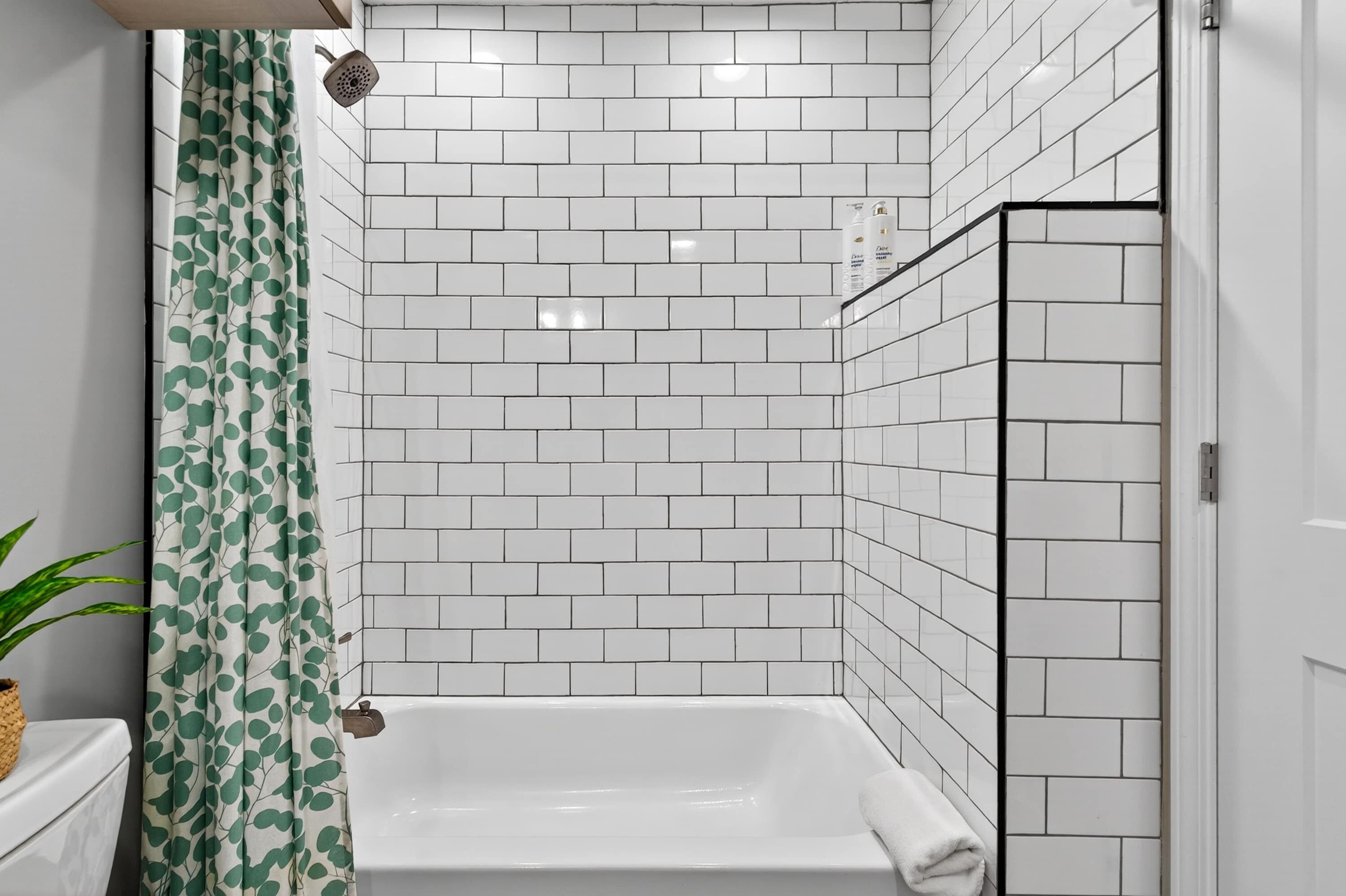 Un baño con paredes de azulejos blancos, una cortina de ducha con estampado de hojas verdes y una bañera blanca.