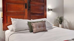 Una cama con ropa de cama blanca, una puerta de madera y una lámpara fijada a la pared.
