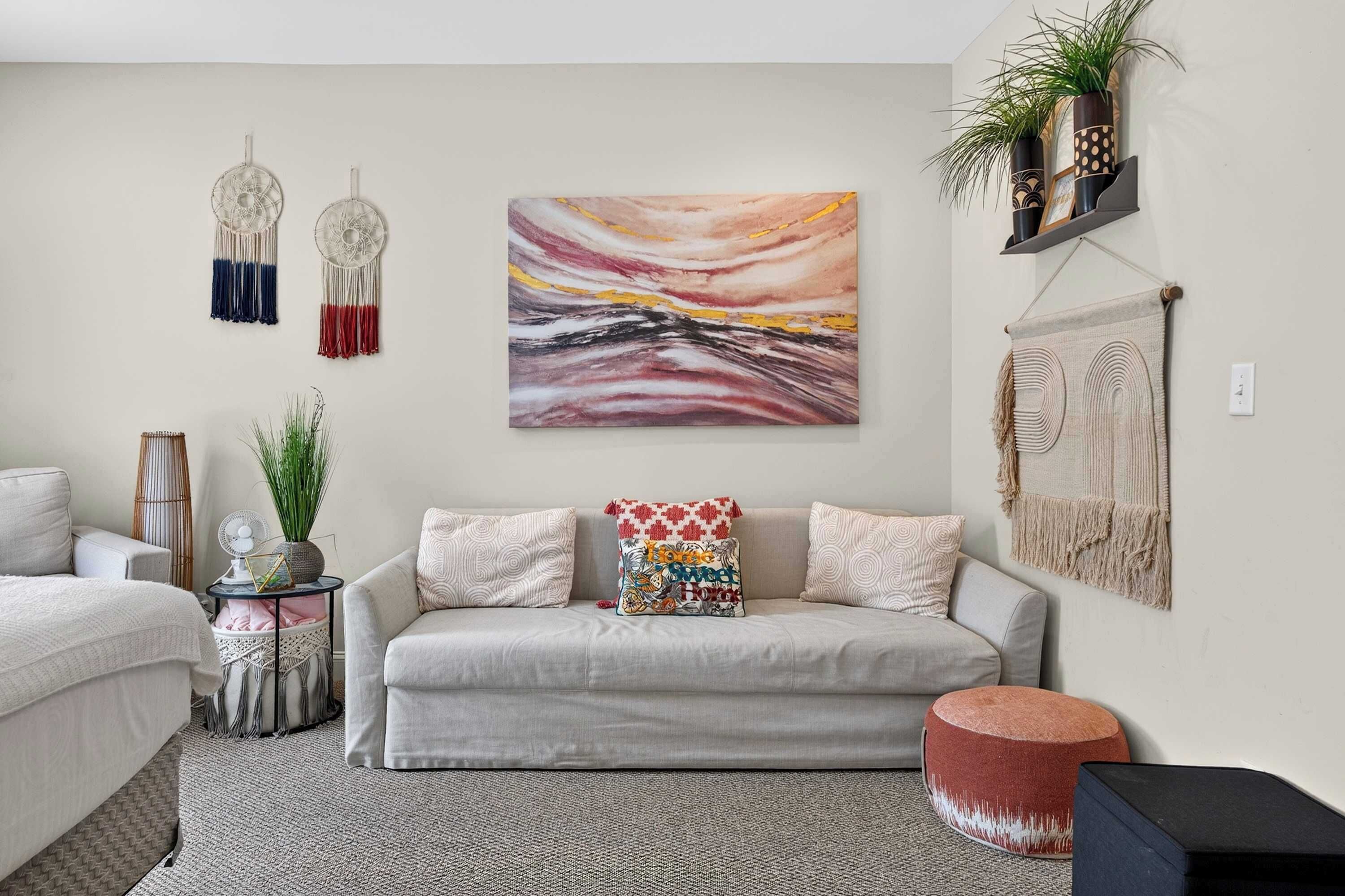 A living room with a sofa, a wall-mounted abstract painting, macramé wall hangings, and a shelf with plants.