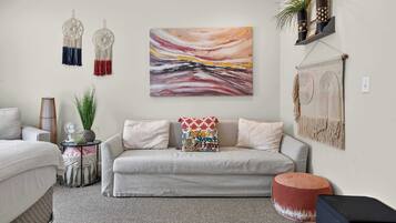 A living room with a sofa, a wall-mounted abstract painting, macramé wall hangings, and a shelf with plants.