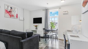 Een moderne, goed verlichte kamer met een grijze bank, een eettafel met stoelen, een aan de muur bevestigde televisie en een raam met gordijnen.