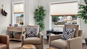 A living room with two beige armchairs, a wooden side table, a large window with blinds, a potted plant, and a wall-mounted shoe rack. - Cozy stay in Germantown near Downtown attractions! (Louisville)