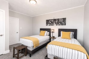 Une chambre avec deux lits, une petite table, une porte et une œuvre d’art accrochée au mur.