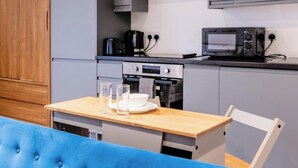Una kitchenette equipada con microondas, cafetera y horno. Hay una mesa con vasos y un plato. Un banco tapizado en azul está en primer plano.