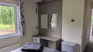 A bedroom with a bed, a bedside table, a mirror, a window with curtains, and a view of greenery outside. - Static Caravan 2 Bed Sleeps 6 Overlooking Water (Northampton)