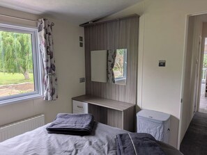 Ein Schlafzimmer mit Bett, Nachttisch, Spiegel, Fenster mit Vorhängen und Blick ins Grüne.