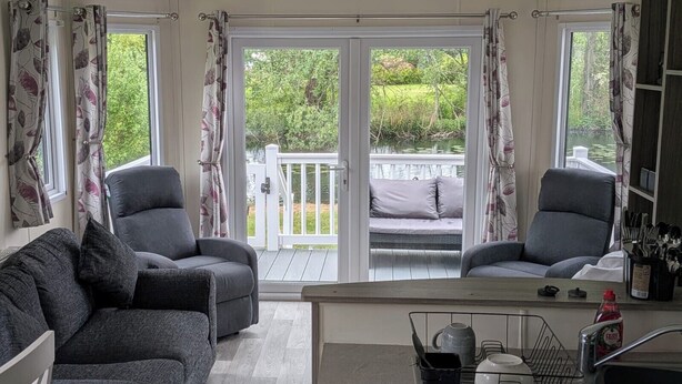 A room with two grey armchairs, a grey sofa, a dining table with a sink, and a view of a deck and greenery outside. - Static Caravan 2 Bed Sleeps 6 Overlooking Water (Northampton)