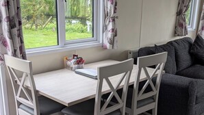 A dining area with a table and chairs, a window with curtains, a radiator, and a grey sofa. - Static Caravan 2 Bed Sleeps 6 Overlooking Water (Northampton)