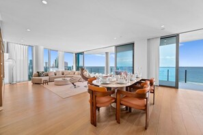 A modern living room with a dining area, large windows, and a view of the ocean. - Signature Oceanview Residence | 2 Car Spaces (Broadbeach)