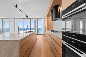 A modern kitchen with a large island, a built-in oven, and a view of the ocean. - Signature Oceanview Residence | 2 Car Spaces (Broadbeach)