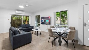 A modern living room with a sofa, a dining table set for four, and a TV mounted on the wall. - Peaceful Retreat Near Shops & Cafés|Secure Parking (Mount Warren Park)